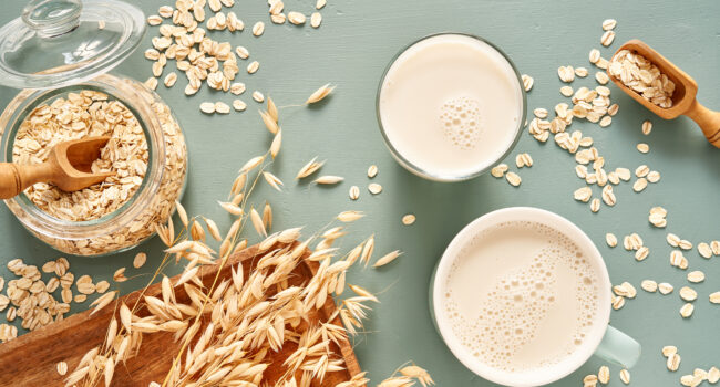 Oat milk in a glass and mug on a blue background. Flakes and ears for oatmeal and granola on a wooden plate.